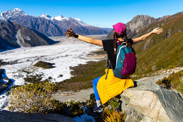 Brave hiker girl sits on rock with arms spread, admiring alpine mountain landscapes and glacial river on trail to Mueller Hut, Aoraki Mount Cook National Park, Canterbury, South Island, New Zealan