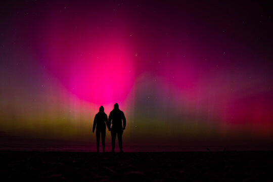 Silhouette of couple holding hands and gazing at sky lit up by vibrant pink and green southern lights, Aurora australis, on beach near Christchurch, Canterbury, South Island, New Zealand