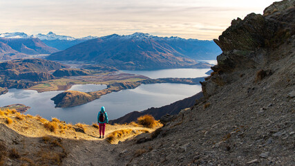Hiker girl hikes along Roys Peak trail and admires stunning panoramic views over Lake Wanaka and surrounding alpine peaks near Wanaka, Otago, South Island, New Zealand.