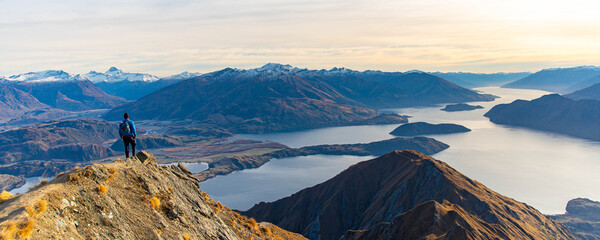 Hiker man stands on summit of Roys Peak and admires stunning panoramic view over Lake Wanaka and surrounding snow covered peaks near Wanaka, Otago, South Island, New Zealand