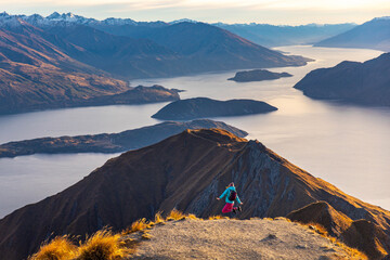 Hiker woman jumps with joy on summit of Roys Peak and admires breathtaking panoramic view over Lake Wanaka and surrounding snow covered peaks near Wanaka, Otago, South Island, New Zealand.
