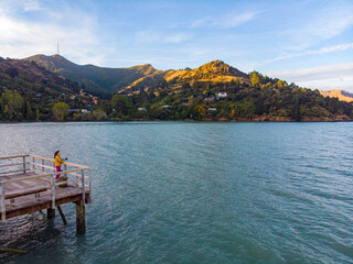 Aerial view of a girl standing at the end of Governors Bay jetty near Lyttelton, Banks Peninsula, South Island, New Zealand, with calm waters and majestic mountainous landscape in the background