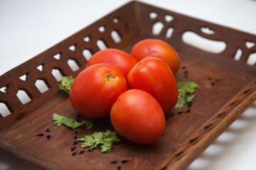 A rustic wooden tray holds a cluster of ripe red tomatoes, their skin glossy with freshness. A few coriander leaves add a subtle touch of green, enhancing the natural appeal of this farm fresh display