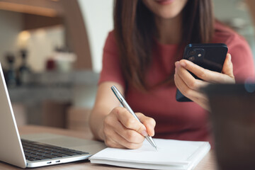 Woman in casual business clothes working on laptop and using smartphone, writing on notebook at...