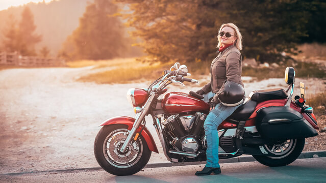 Confident woman with sunglasses posing with red cruiser motorcycle on a scenic countryside road at sunset, holding helmet and smiling proudly.