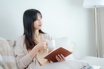 A young Asian woman is comfortably nestled on a sofa at home, engrossed in reading a book while...