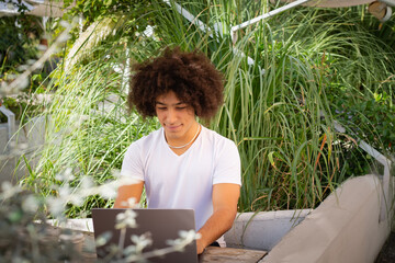 Smiling young ethnic guy , dressed in casual clothes, works on a laptop outdoors, in a green area in nature. Green open space for coworking, freelancer, relaxing, coffee, food. Free place to work
