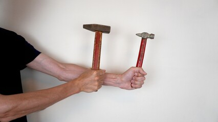 A man holds two different hammers with red handles on a light background. One hammer has a wide...