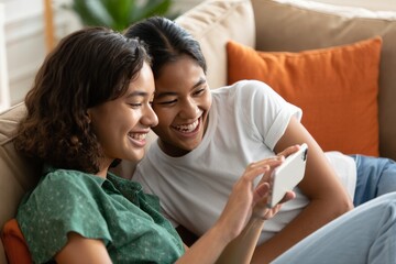 Two friends laughing together watching a phone screen on the couch at home indoors