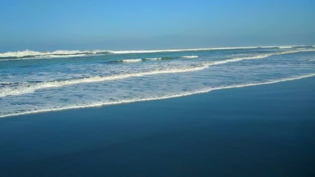 Gentle waves roll onto the sandy shore under a clear blue sky, creating a calm and serene coastal atmosphere.