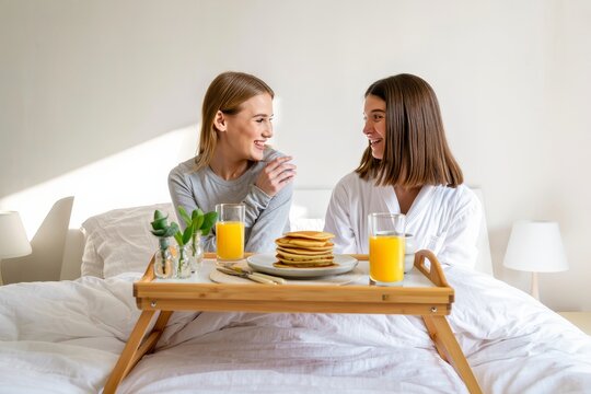 Two women enjoying breakfast in bed with pancakes and orange juice in the morning light - Powered by Adobe
