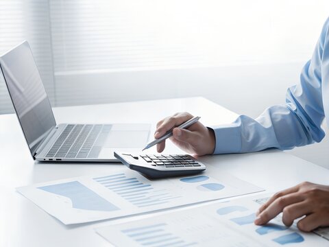 A persons hands operate a calculator and pen over financial documents with a laptop in the background