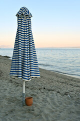 A closed blue-and-white striped beach umbrella stands in the sand near the shoreline, with gentle sea waves and a soft pastel sunset sky in the background. A small bucket lies nearby, adding a touch o