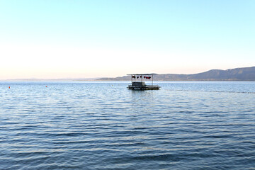 A peaceful scene of a small boat floating on a calm sea, with gentle waves and distant mountains under a soft blue sky. The tranquil water and minimalistic composition create a serene and relaxing atm