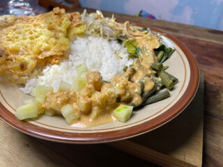 pecel is a stew of long beans, sprouts and mustard greens sprinkled with crushed peanut sauce and peanut brittle as a complement, a close up of warm pecel rice served on a ceramic plate on the table