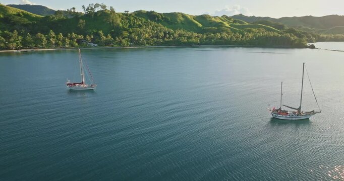 Fiji islands: Two sailboats mooring calm ocean water near tropical coast covered lush vegetation, some traditional fijian houses during a sunny day. Wild nature travel landscape. Aerial drone flight