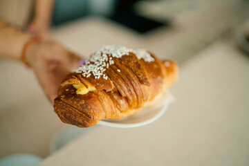 Delicious Freshly Baked Golden Brown Croissant Topped with Sugar Sprinkles Served on a White Plate with a Hand Holding it in a Cafe Setting