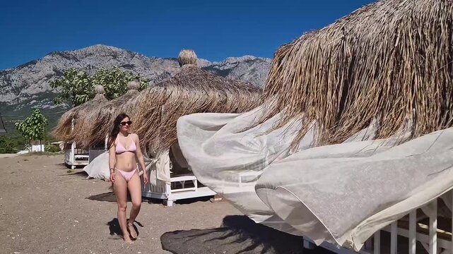 Sexy young woman in bikini and sunglasses confidently walks on a sandy beach with rustic thatched huts. Majestic Taurus mountains rise in the background under a clear blue summer sky in Kemer Turkey