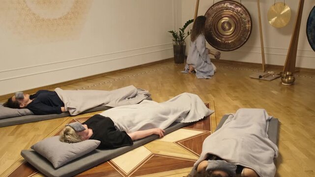 Participants lie on mats with eye masks while a female sound therapist plays a ceremonial gong during a guided group sound meditation session focused on healing and relaxation