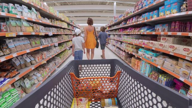 Family shopping in supermarket, viewed from shopping cart perspective. Mother with two kids walking down aisle filled with food products and groceries