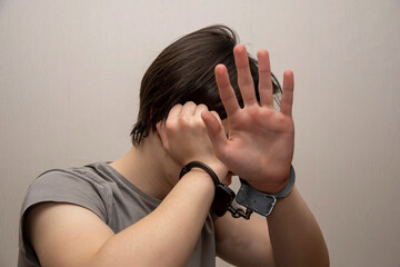 A handcuffed teenager covers his face with his hands on a gray background, medium plan. Juvenile...