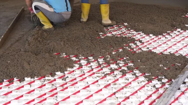 Workers pour concrete mortar onto the top of underfloor heating pipes during construction.