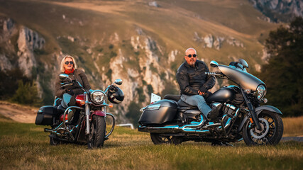 Stylish mature couple sitting on cruiser motorcycles in scenic mountain landscape, enjoying the moment during a peaceful road trip at sunset.