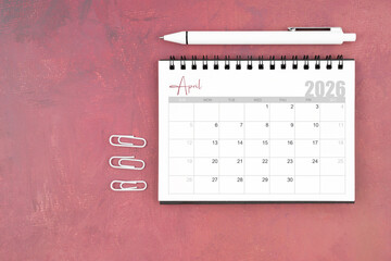 Desk calendar for April 2026 and pen with paper clips on red table, Flat lay.