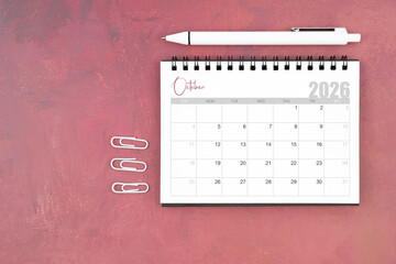 Desk calendar for October 2026 and pen with paper clips on red table, Flat lay.