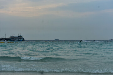 Busy ocean scene with a pier, ferry, jet skis, and distant cargo ship in Thailand. 