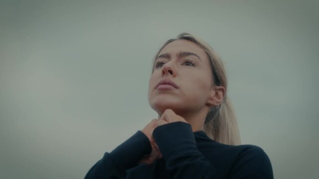 A female in athletic clothing, zipping up near the beach or sea under an overcast sky.