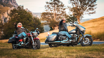 Cheerful biker couple pointing toward the camera while sitting on cruiser motorcycles in golden...