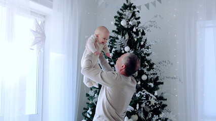 Grandfather lifting baby grandson in front of a festive christmas tree, creating a heartwarming holiday moment. A day with family before Christmas. New Year's Eve with family