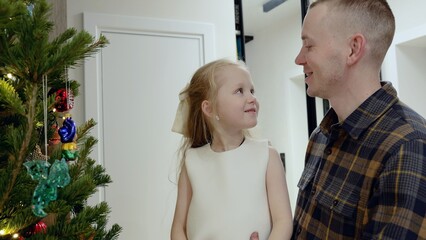 Father helping young daughter hanging ornaments while decorating festive christmas tree, sharing tender moment at home during holiday celebration