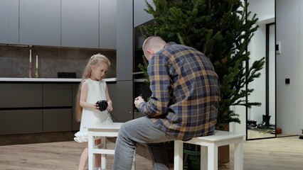 Father helping young daughter untangling tangled christmas lights, preparing holiday decorations in bright, contemporary kitchen with warm, festive mood