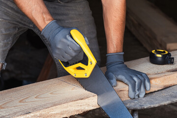 Brutal carpenter saws board with hacksaw in gray gloves.