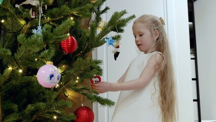 Young blond girl carefully hanging colorful ornaments on decorated christmas tree, creating magical holiday ambiance with twinkling lights and festive charm