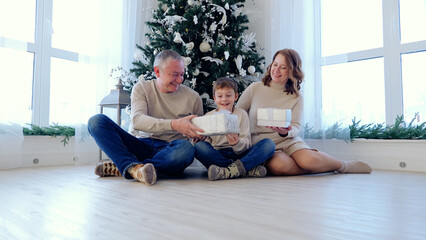 Young boy receiving christmas presents from his happy parents, sitting on the floor in a bright living room with a decorated christmas tree. A day with family before Christmas. 