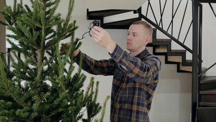 Young male hanging sparkling garland lights on elegant christmas tree, creating festive winter holiday ambiance inside contemporary home setting