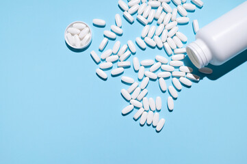 White container, plastic packaging and scattered white tablets, vitamins, pills, food supplements on blue background top view. Medical and pharmaceutical concept, health concept