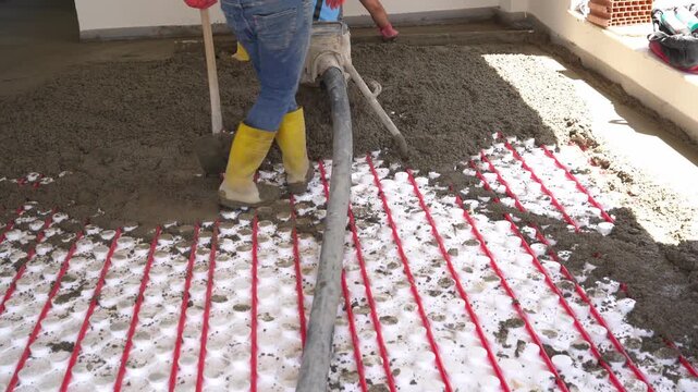 Workers pour concrete mortar onto the top of underfloor heating pipes during construction.