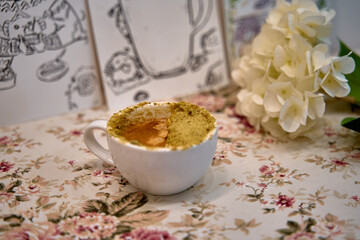 A Delicate Cup of Coffee Topped with a Beautiful Layer of Sweet Cream and adorned by Fresh Flowers on a Soft Floral Tablecloth