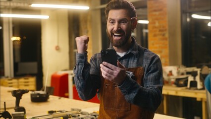 Happy Caucasian winner man guy wood master win scrolling mobile phone in workshop carpenter...