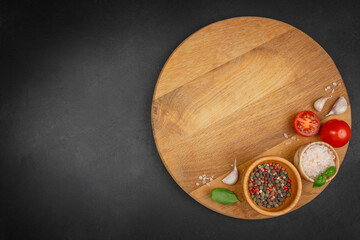 Top view of wooden round board with salt, pepper, tomatoes, garlic, and basil on black background....