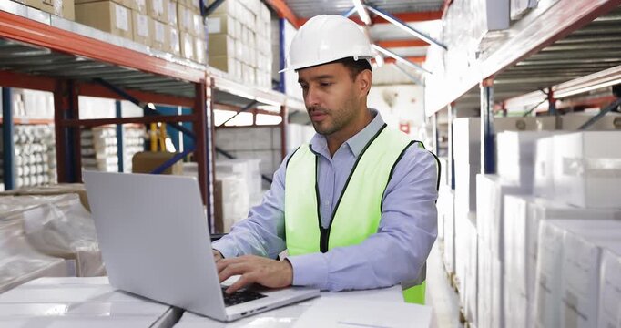Young Hispanic warehouseman worker wearing safety vest and helmet working in warehouse using laptop, reviewing paper documents, preparing report, controlling inventory, engaged in logistics process