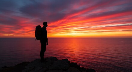 Fototapeta premium A silhouetted hiker watches a fiery sunset over the ocean horizon, a moment of peace.