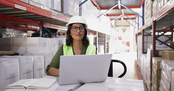 Female warehouse worker wearing hardhat and vest working on laptop, check notes in planner next to large stacks of boxes on shelves. Logistics coordination, inventory tracking, supply chain management