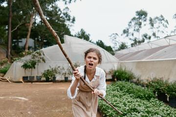 Young woman expressing frustration while working in a greenhouse, holding a stick amidst the...