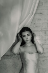 Fototapeta premium A young woman wearing a white corset dress standing against a textured wall with hands near her head