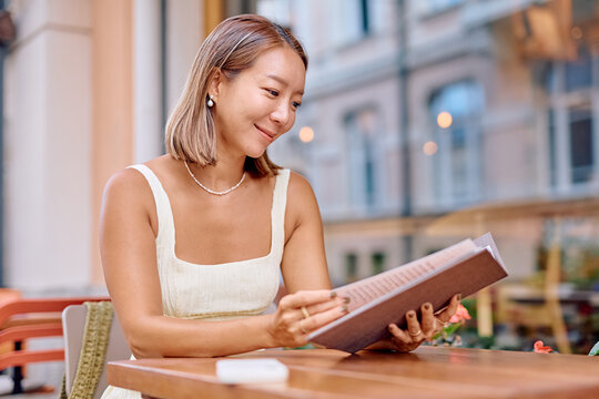 Happy woman sitting at a table on a summer terrace, enjoying the experience of choosing a meal from the menu at a cafe